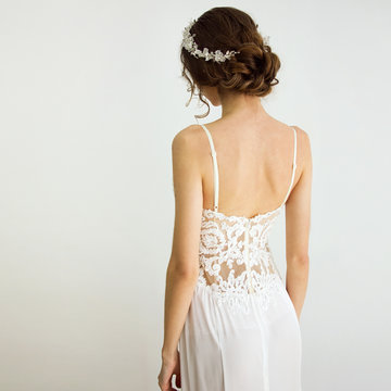young brunette sexy girl in a white wedding boudoir dress and jewellery in hairstyle is standing from the back  like swan princess on a white wall background 