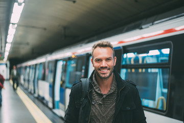 Happy young blonde man with backpacking inside the underground station waiting for the train