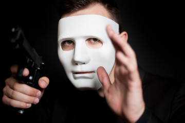 Close-up of a man in a white mask on a black background. Holds the gun with his arms raised