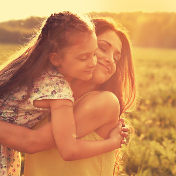 Happy enjoying mother strong hugging her relaxing kid girl on sunset bright summer background with closed eyes. Closeup toned orange color portrait of family love.