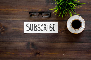 Email subscribe concept. Hand lettering subcribe on work desk with plant, glasses, cup of coffee on dark wooden background top view copy space