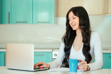 beautiful young woman student (freelancer) in a white dress and black hair working at home with a laptop in a turquoise color kitchen. In the hands of a cup, the idea of happiness