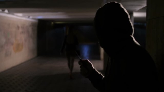 Woman walking alone in dark underpass at night, male silhoette in hood appears, man's hand opening folding knife with blade and criminal starts following his prey. Killer going to attack the victim