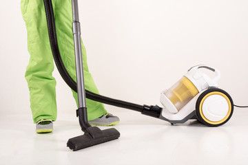 Happy guy in bright green pants and hat removes the dust in house with the help of yellow compact vacuum cleaner.