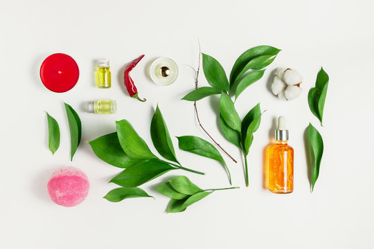 Flatlay of serum, perfume, bath bomb, essential oils with ruscus leaves, pepper and cotton flower on white as a concept of natural body and skincare