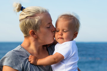 Fototapeta premium Young hipster mother kisses her blonde baby with closed eyes. Portrait of mom and child against blue sea. Motherhood, mother and baby relationship and tenderness concept