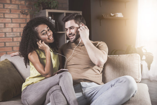 Having fun together. Portrait of delighted young boyfriend and girlfriend are sitting on sofa and listening to music on smartphone. They are using headphones. Man is looking at girl with adoration