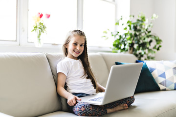 Happy little girl at home working with a laptop on sofa