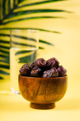 Wooden bowl of dates fruits with glass of water for iftar in ramadan fasting month, green palm tree leaf branch on yellow background, copy space. Eid mubarak celebration