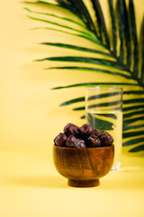 Wooden bowl of dates fruits with glass of water for iftar in ramadan fasting month, green palm tree leaf  branch on yellow background, copy space. Eid mubarak celebration 