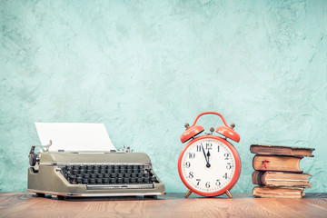 Retro classic typewriter with sheet of paper, old books and big orange alarm clock on wooden table front aquamarine concrete wall background. Vintage style filtered photo