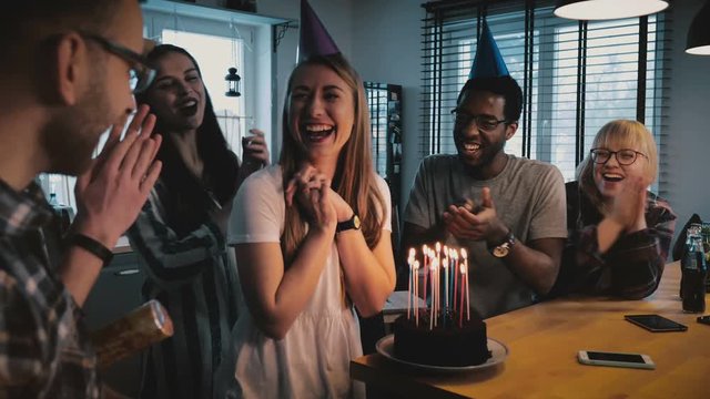 Happy Caucasian girl makes a wish at birthday cake with candles. Multiethnic friends at surprise party slow motion.