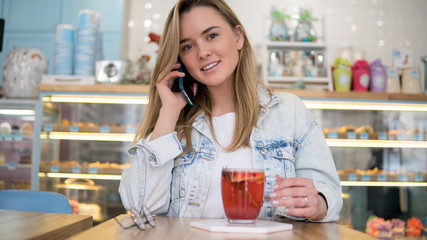 Blond woman drinks tea indoors cafe, smiling and speaking on the phone