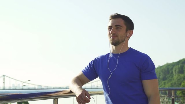 Low angle shot of a handsome bearded athletic man smiling, looking away, relaxing after morning workout outdoors in the city. Happy male athlete listening to music after exercising.