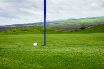 Obraz premium Close up side view of a golf ball on a putting green next to the hole with a blue pin, and hills and a stormy sky in the background 