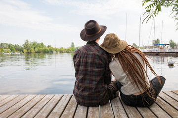 Young couple of travelers sitting on the wooden pier near river, two hipsters with hats and...