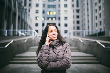 young girl talking on mobile phone in courtyard business center. girl with long dark hair dressed in winter jacket in cold weather speaks on phone on background buildings made of glass and concrete