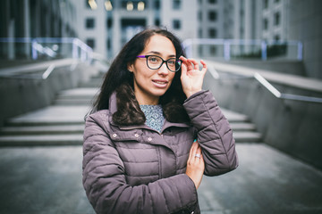 Portrait beautiful young business woman brunette in jacket and sweater stands on background office building, business center in glasses for view. Hand holding onto shackle of glasses and looking away