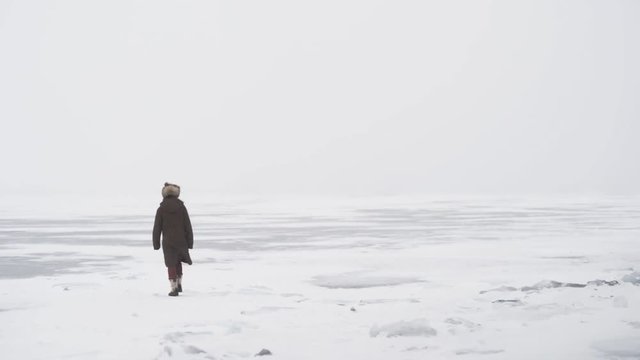 A lonely man in a fur hat walks on a snowstorm on the ice. White snowflakes lay around. Nothing is visible on the horizon, as a snowstorm begins and the weather conditions deteriorate. A man with diny