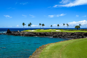 Sunny day on a tropical golf course fairway with the putting green in the distance surrounded by palm trees and sand traps, lava rock, blue pacific ocean, and blue sky and white clouds in background - obrazy, fototapety, plakaty