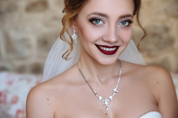 A beautiful bride and a dress with open shoulders. A close-up shot of a girl with a delicate eye makeup and red lips smiles. Light curls of hair, earrings and a thin necklace emphasize the neck