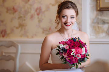 The bride is holding a bouquet of red flowers and smiling happily sitting at the table. A girl with blue eyes and curls gets married.