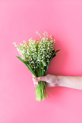 Give a bouquet of delicate lily of the valley flowers. Hand hold bunch of flowers on pastel pink background top view copy space
