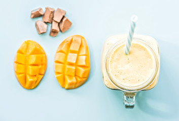 summer drink mango milkshake in mason jar with drinking straw decorated with a half of mango, chocolate pieces flat lay top view on blue background