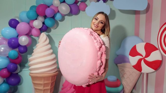 A slender beautiful girl poses in the Studio with a big marshmallow in her hands. Happy Barbie girl with a big marshmallow on a background of balloons and a huge candy on stick.