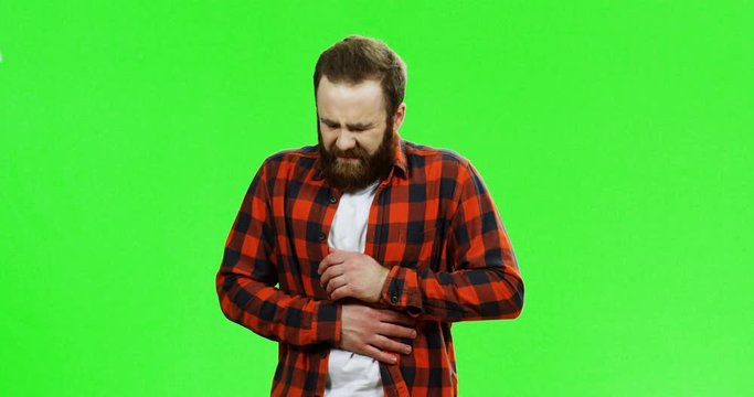 Caucasian man with a beard standing on the green screen background and holding for his stomach as having a strong spasm pain. Green screen. Chroma key.