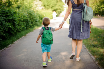 Back view of mother walking down the street with a little son with a backpack on sunny day