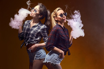 Side view of trendy young female in glasses and shorts standing and smoking on studio background