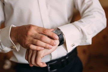 The groom in a white shirt and trousers looks at the wristwatch