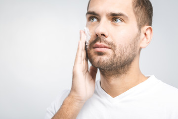 Fototapeta premium Skin care. Handsome young shirtless man applying cream at his face and looking at himself with smile while standing over gray background. Close-Up. Space for text.