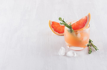 Refreshing cold citrus cocktail with ice cubes, green rosemary and slice grapefruit on soft light white wood background.
