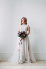 Fototapeta premium Beautiful bride in a wedding dress in a bright stylish Studio. Wedding in European style. Decor and bouquet in hand. Young girl standing, sitting and posing