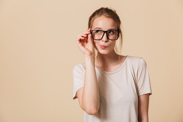Picture of charming blond woman 20s wearing basic t-shirt touching eyeglasses with flirty look, isolated over beige background in studio - obrazy, fototapety, plakaty
