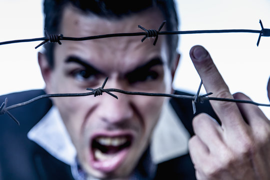 Fuck the system concept. Man behind a barbed wire shows a middle finger gesture as symbol of lack of freedom