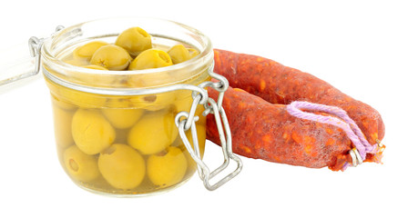 Glass storage jar filled with green olives next to a whole chorizo salami isolated on a white background