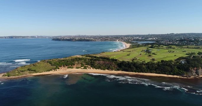 Waterfront of Long Reef headland on Sydney’s northern beaches approached from open sea in aerial flying on a sunny day.
