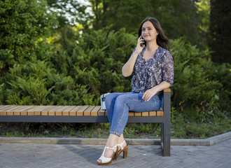 Girl sitting on bench and talking on phone. Woman in the park on summer day