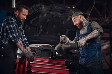 Fototapeta na wymiar Two bearded auto mechanic in a uniform and safety glasses working with an angle grinder while standing against a broken car in repair garage. 