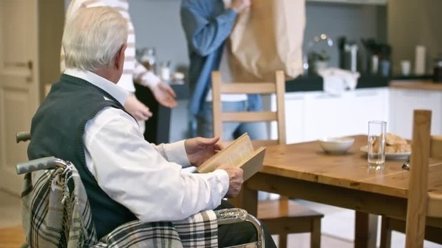 Tilt up of cheerful female caregiver walking into kitchen and chatting with elderly man in wheelchair while male volunteer unpacking groceries