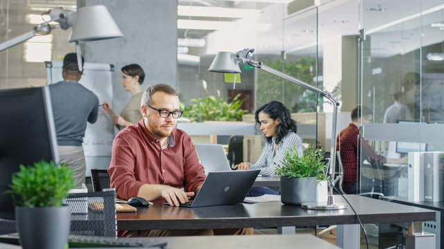 Busy International Corporate Office, Caucasian Man Working at His Desk on a Laptop. In Background Creative Young People Working.