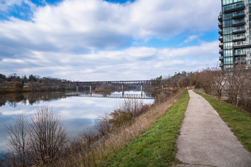 Narrow Path alongside a River and Blue Sky with Clouds on a Late Autumn Day. An Old  Railway Bridge Crossing the River in Visible in Background. Reflection in Water.
