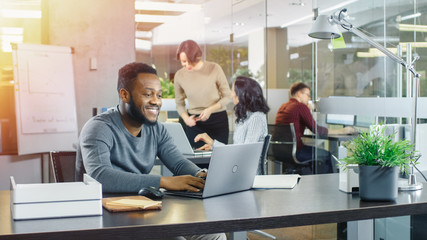 Busy International Office, African-American Man Working at His Desk on a Laptop, in the Background Businesswomen Discuss Relevant Data. Stylish Office with Talented Young People.