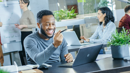 Man Working at His Desk Working on a Laptop, Encounters Funny Mistake and Laughs. In the Background Creative Young People Doing Their Jobs. Modern Bright Office.