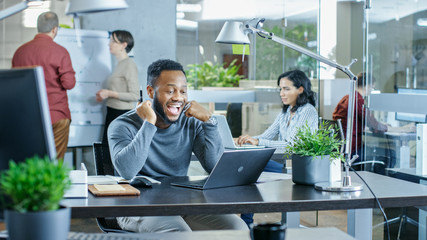 Man Working at His Desk Working on a Laptop, Encounters Funny Mistake and Laughs. In the Background Creative Young People Doing Their Jobs. Modern Bright Office.