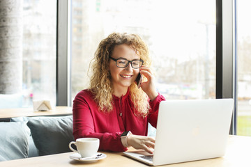 Attractive young woman sitting at coffee shop with her fashionable laptop, drinking cappuccino, talking on the phone, receiving good news smiling. Beautiful blonde female with curly hair. Background.