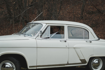 Just married wedding couple is sitting in the retro vintage car in the park. Spring or autumn grey day in forest. Blonde bride in elegant white dress  and elegant groom in love are smiling.